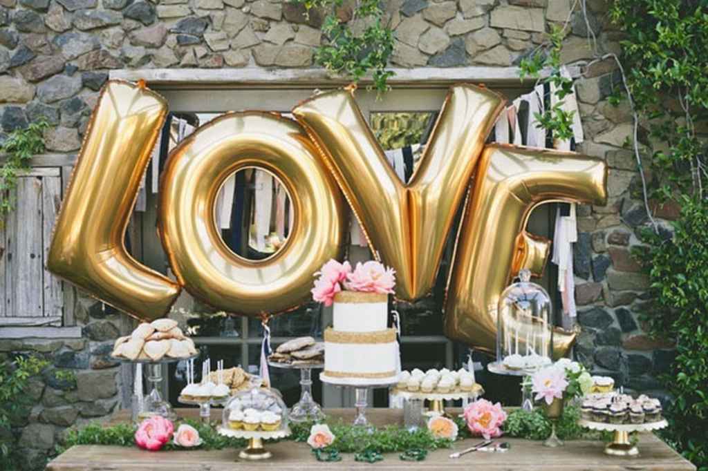 Zlat Love napis iz balonov je ozadje elegantne sladke mize na kateri so raznovrstne sladice in tudi poročna torta, ki je okrašena s cvetjem. Foto: One love photography