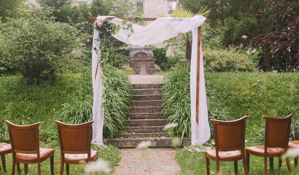 Rustikalno obarvan civilni poročni obred postavljen v vrtu gradu Brdo. Lesen obok za poroko je okrašen z belim blagom, cvetjem ter lesenimi vintage stoli na levi in desni strani oboka. Foto: Nastja Kovacec Photography