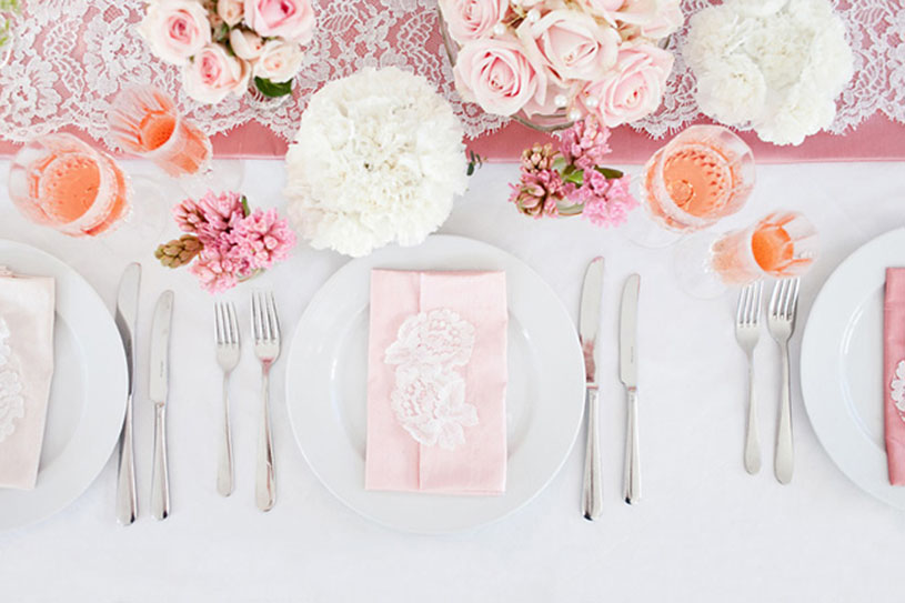 Na mizi z belim prtom so postavljeni krožniki, kozarci, prtički ter cvetlična poročna dekoracija v roza barvi. Foto: vir Belle the magazin