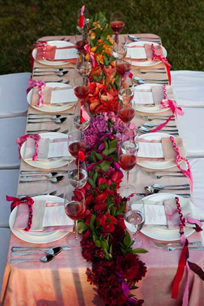 Cvetlična dekoracija poročnega omizja, ki se preliva iz bordo rdeče v rumeno barvo. Poleg cvetlične dekoracije je tudi prt v prelivajoči barvi, s tem, da je rdeča pri vznožju mize. Foto: vir La Pertie Events