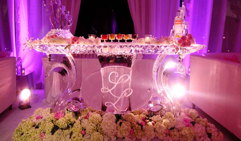 Na roza osvetljenem šanku iz ledu so postavljene alkoholne pijače. Pred njim pa stoji prekrasna cvetlična dekoracija. Foto: vir Ice Scoulpture