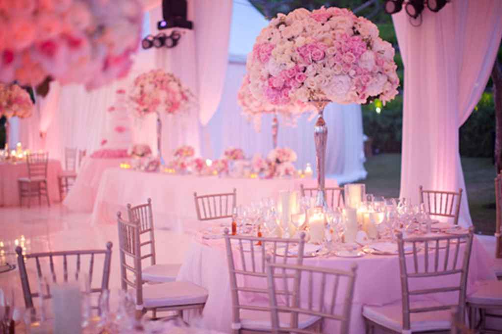 Elegantna poročna dvorana v roza svetlobi. Na mizah so postavljeni visoki poročni cvetlični aranžmaji, ki so v odtenkih roza in bele pod njimi pa nešteto svečk različnih višin v kozarcih. Foto: Samuel Lippke