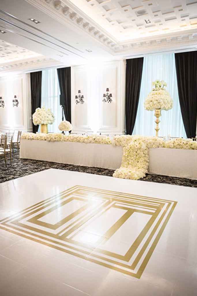 Moderna poročna dekoracija v belo, črnih in zlatih odtenkih. Na glavni mizi se vije dolga girlanda iz belih vrtnic, na oknih so črne zavese ter na plesišču narisan logotip v zlati barvi. Foto: Ikonica