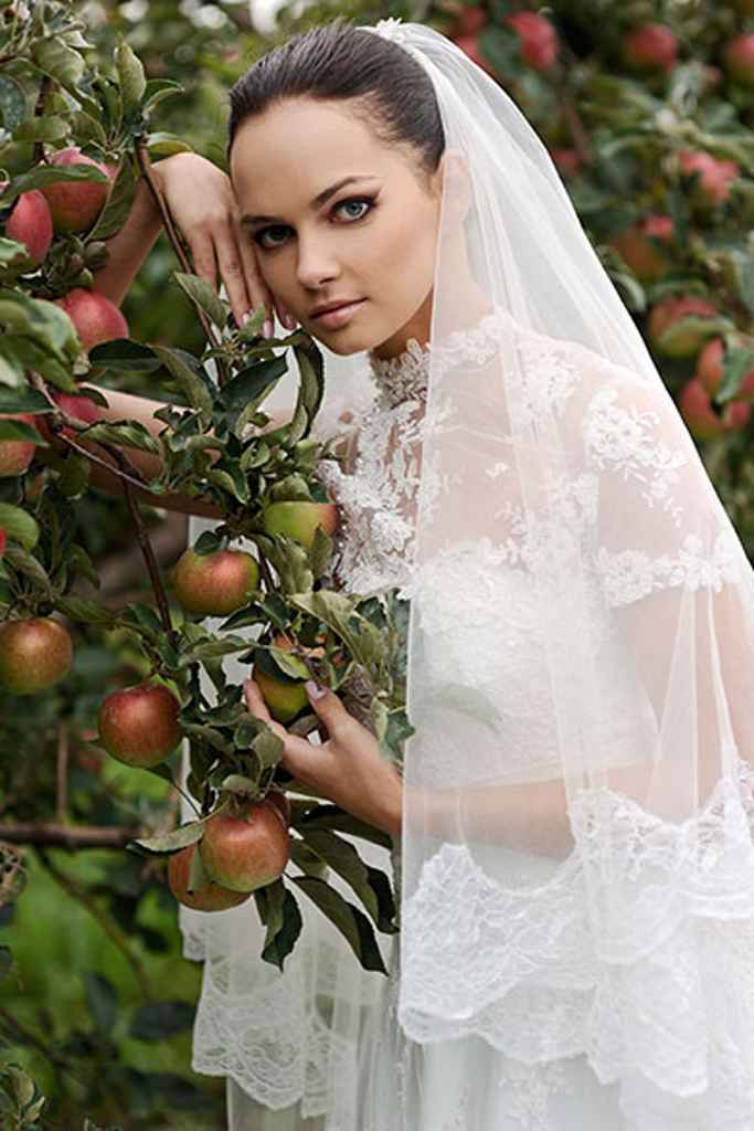 Prekrasna nevesta v beli čipkasti poročni obleki s pajčolanom s čipkastim robom je naslonjena na jablano. Foto: arhiv Storija weddings