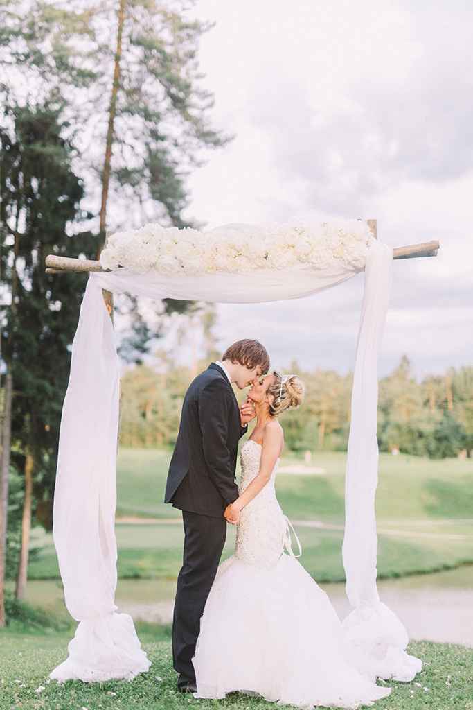 Nevesta in ženin se poljubljata pod cvetličnim obokom iz belih vrtnic in belega blaga. Foto: Nastja Kovacec Photography