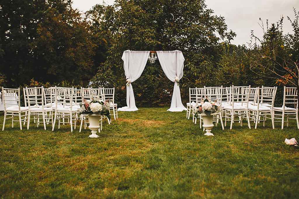 Prekrasna postavitev civilnega obreda na prostem z belimi stoli, lesenim cvetličnim obokom in prekrasnim cvetjem v pastelnih tonih. Foto: Aleks & Irena Kus Wedding Photography