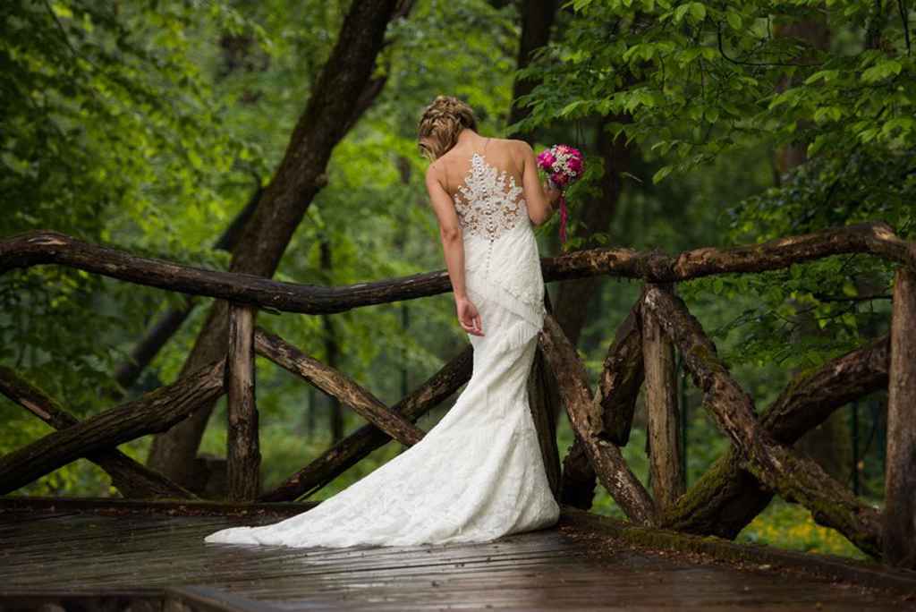 Nevesta v prečudoviti poročni obleki s čipkastim hrbtnim delom in vlečko hodi po mokrem mostu v Arboretumu. Foto: Jean Kanoyev Photography