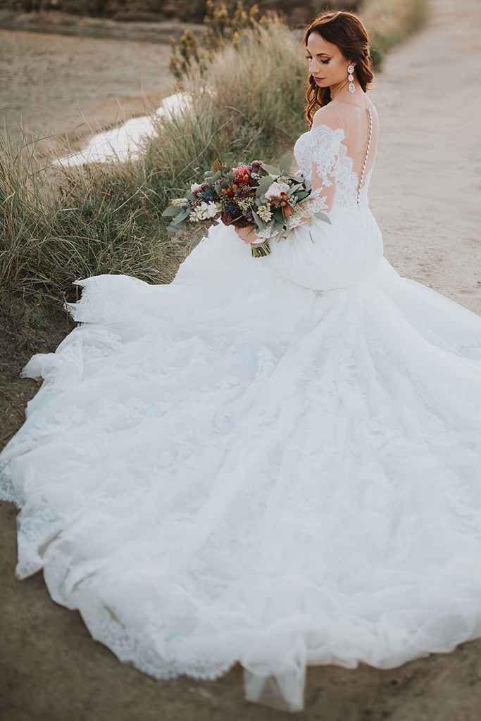 Prekrasna nevesta v poročni obleki z dolgo vlečko in dolgimi rokavi kleči na Strunjanski obali in pozira fotografinji. Foto: Ana Gregorič Photography 