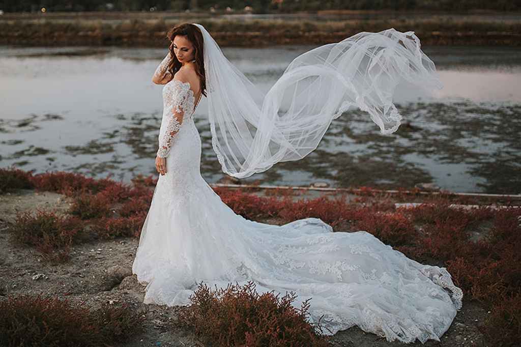 Nevesta se fotografira v svoji poročni obleki na plaži med tem, ko ji veter v zrak dvigne njen pajčolan. Foto: Ana Gregorič Photography