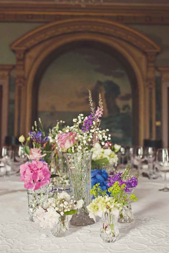 Razgibana poročna cvetlična dekoracija v modri, roza, zeleni in beli barvi, ki je postavljena na sredino mize z belim prtom. Foto: Joanna Brown Photography