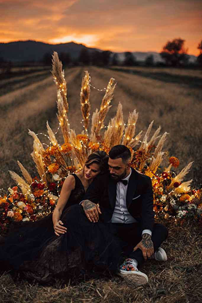 Zaročen par med sončnim zahodom sedi na tleh pred boho ležečim cvetličnim obokom, ki ga krasijo novoletne lučke. Foto: JS Oblikovanje